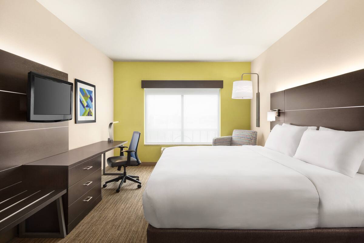 Habitación de hotel con cama, escritorio, silla, televisor y ventana con persianas.