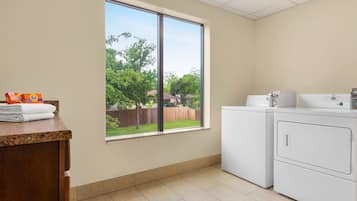 Une buanderie avec une grande fenêtre, un lave-linge et un plan de travail sur lequel sont pliées des serviettes et où se trouve du détergent à lessive.