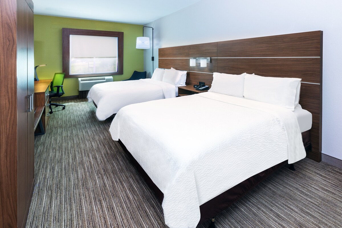 A hotel room with two beds, a desk with a chair, a lamp, and a window with blinds.
