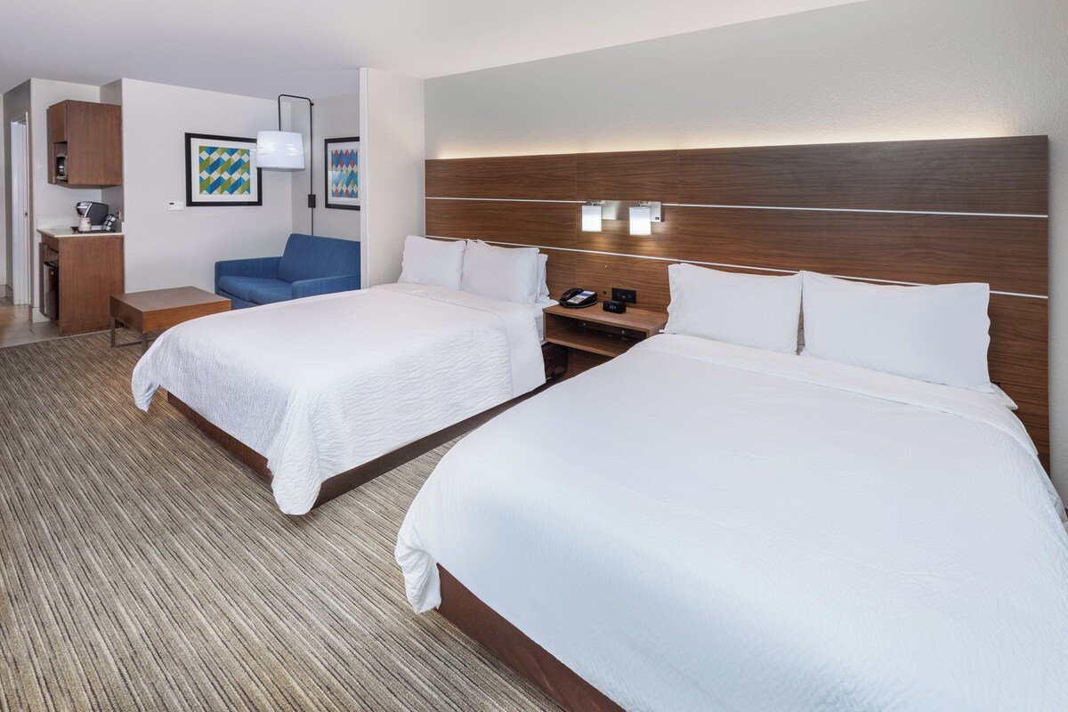 A hotel room with two beds, a desk, a chair, and a small kitchenette.
