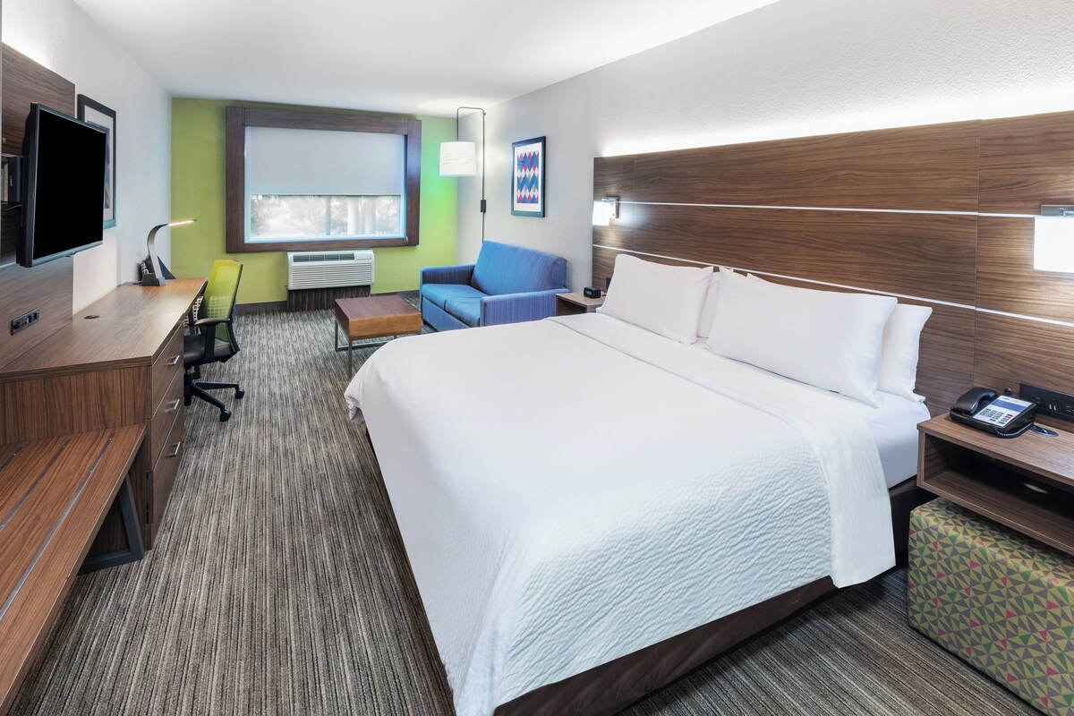 A hotel room with a large bed, a desk with a chair, a TV, and a window with blinds.