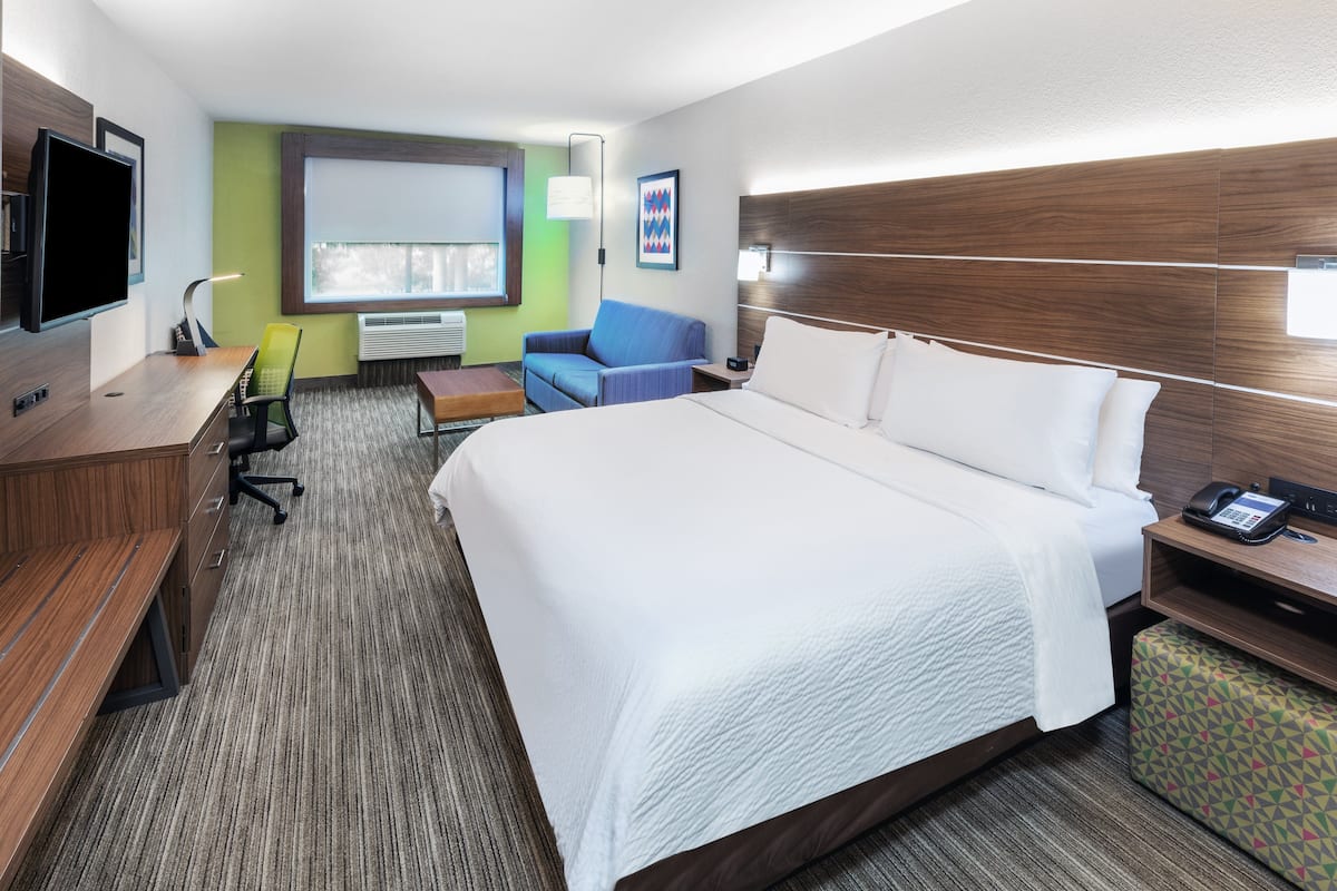 A hotel room with a large bed, a desk with a chair, a TV, and a window with blinds.