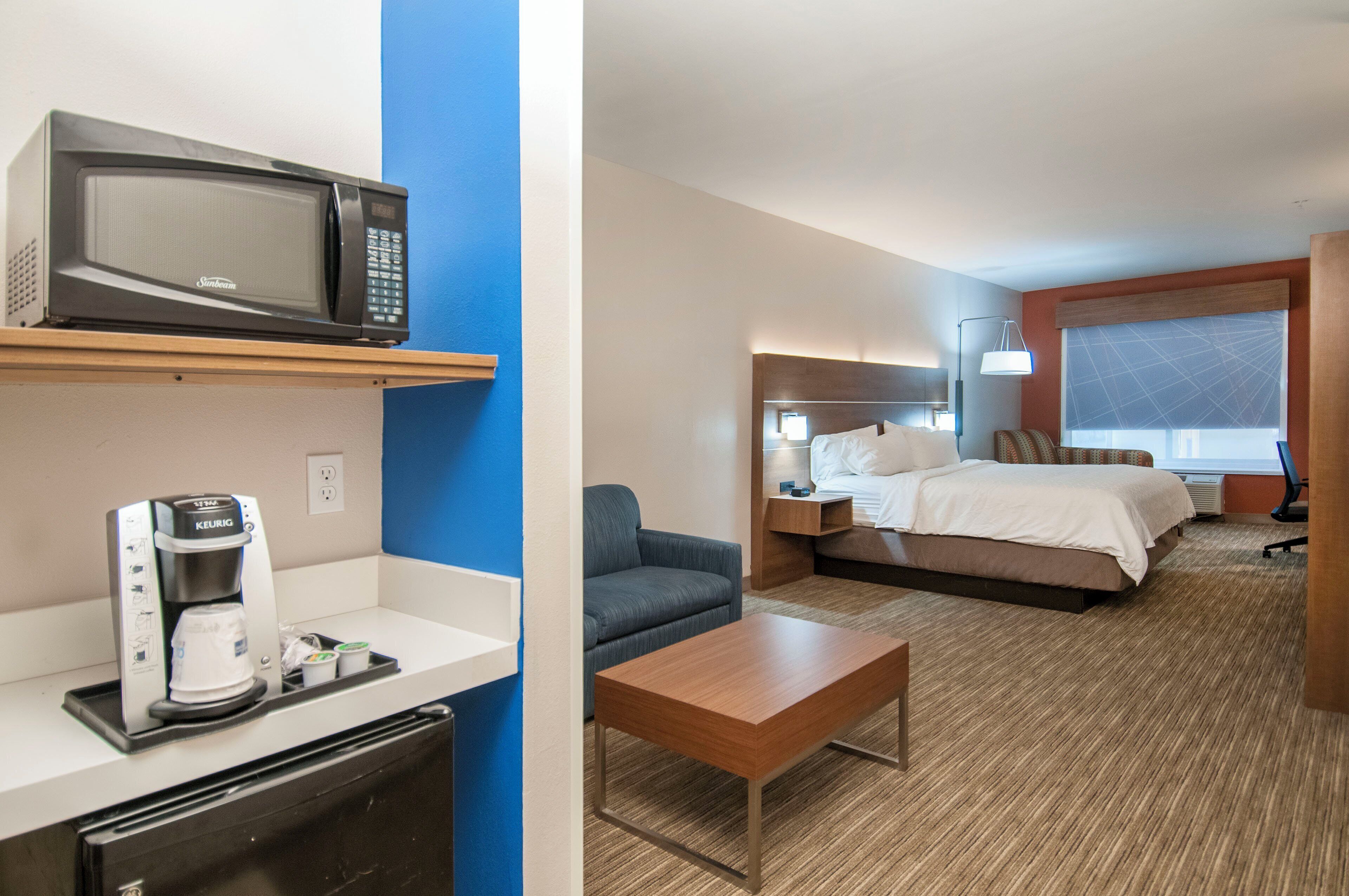 Habitación de hotel con cama, mesita de noche, lámpara y una pequeña cocina equipada con microondas y cafetera.