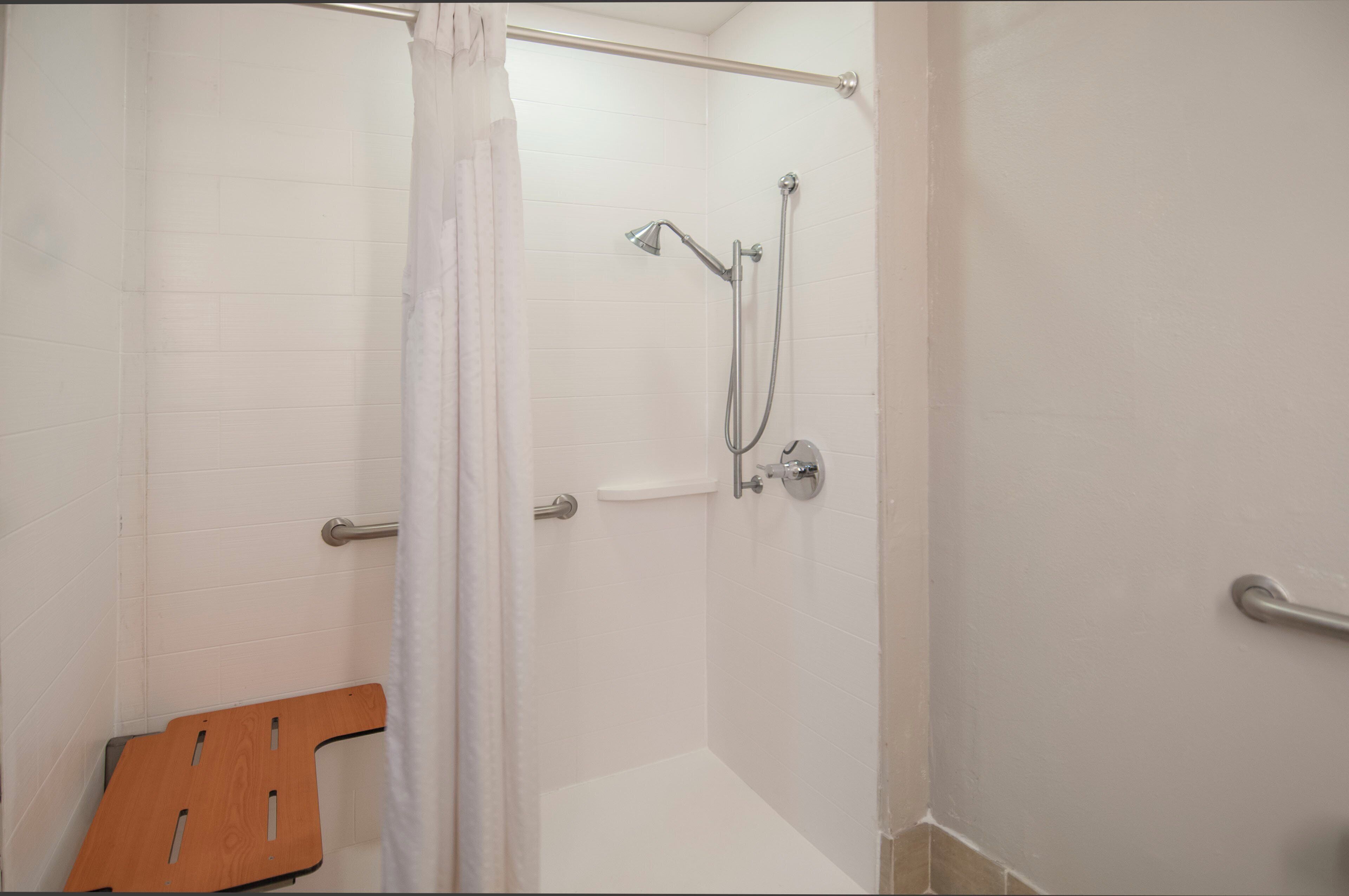A shower area with a white tiled wall, a showerhead, a handheld shower attachment, and a wooden bench.