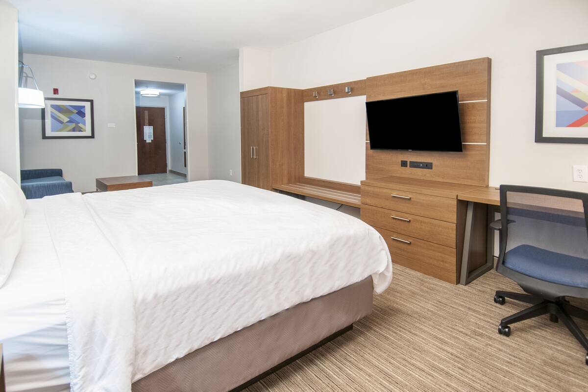 A hotel room with a large bed, a desk with a chair, a TV mounted on the wall, and a framed picture on the wall.