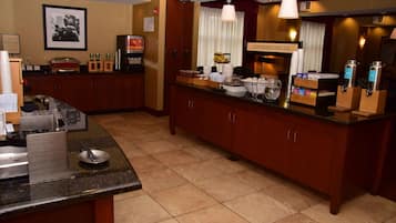 A hotel breakfast area with a buffet setup, including a coffee machine, food containers, and a sign indicating breakfast is served here.