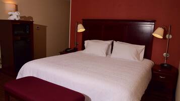 A hotel room with a large bed, two bedside lamps, a wooden nightstand, a small desk with a lamp, and a red bench.