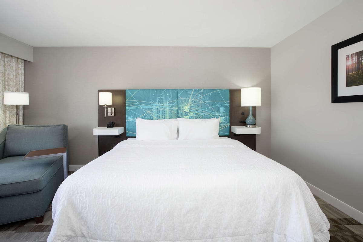 A hotel room with a large bed, a blue abstract wall art, two bedside lamps, a side table, and a blue armchair.