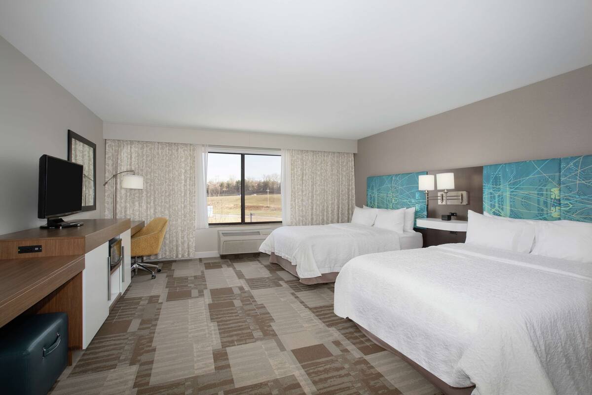 A hotel room with two beds, a desk with a television, a chair, and a large window.