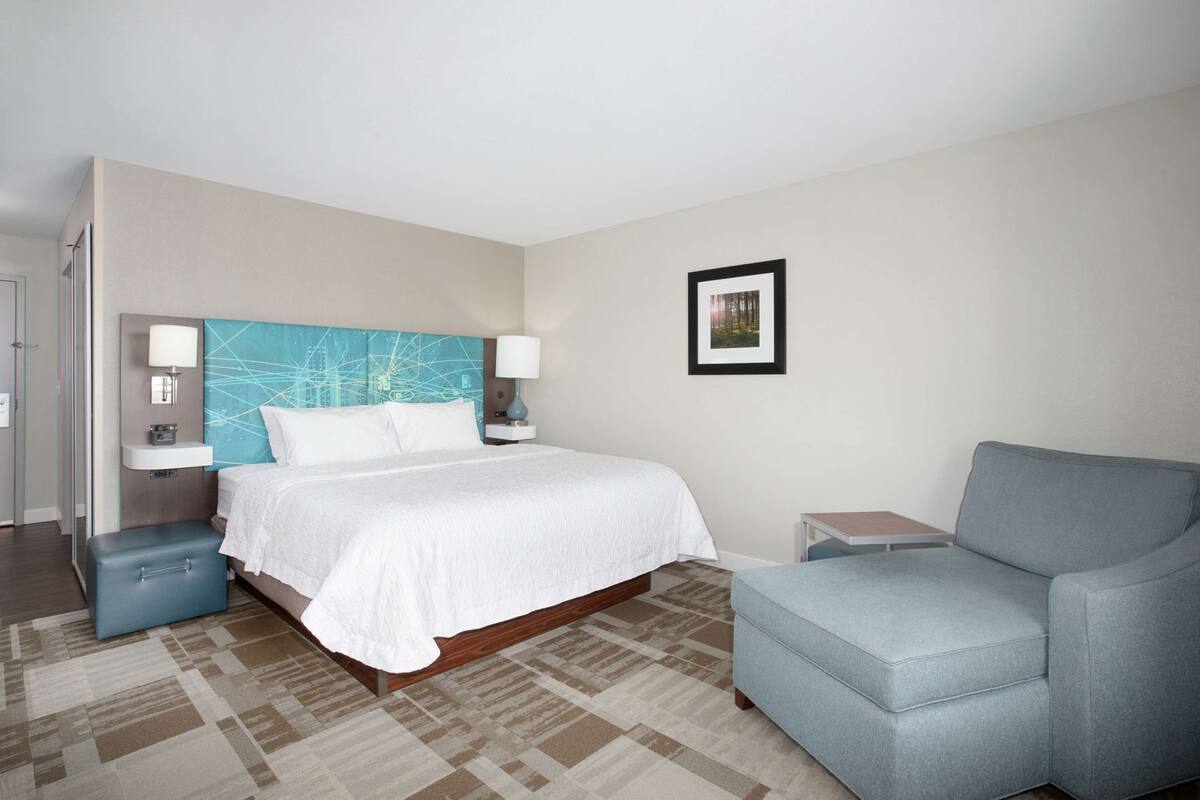 A hotel room with a bed, a gray armchair, a nightstand, a wall-mounted picture, and a window.