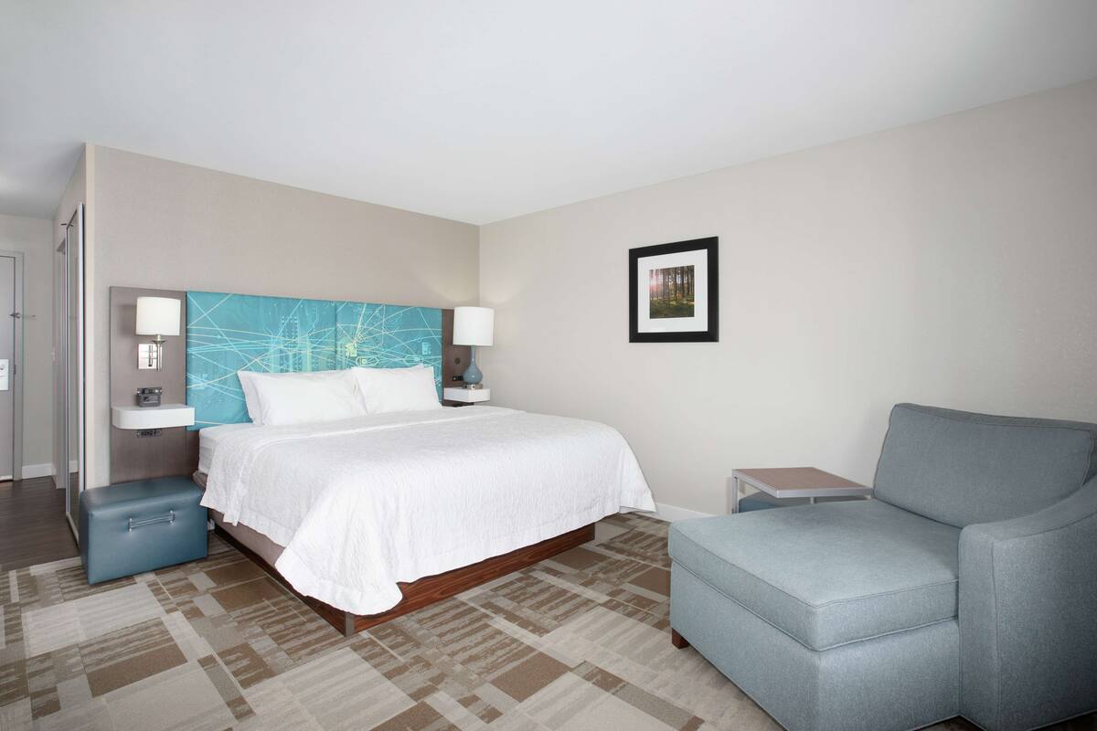 A hotel room with a bed, a blue armchair, a small table, and a framed picture on the wall.