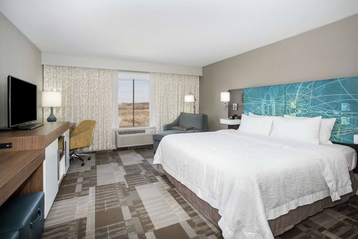 A hotel room with a bed, a desk with a computer, a chair, a window with curtains, and a large abstract painting on the wall.