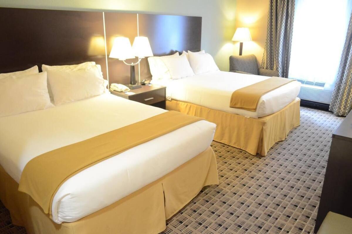 A hotel room with two beds, each with white linens and a beige throw blanket. There are bedside lamps and a nightstand with a phone. The room has a patterned carpet and a window with curtains.