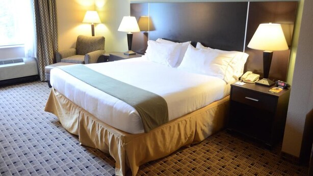 Une chambre d’hôtel avec un grand lit, deux lampes de chevet, une chaise, une table de chevet avec un téléphone et une fenêtre avec des rideaux.