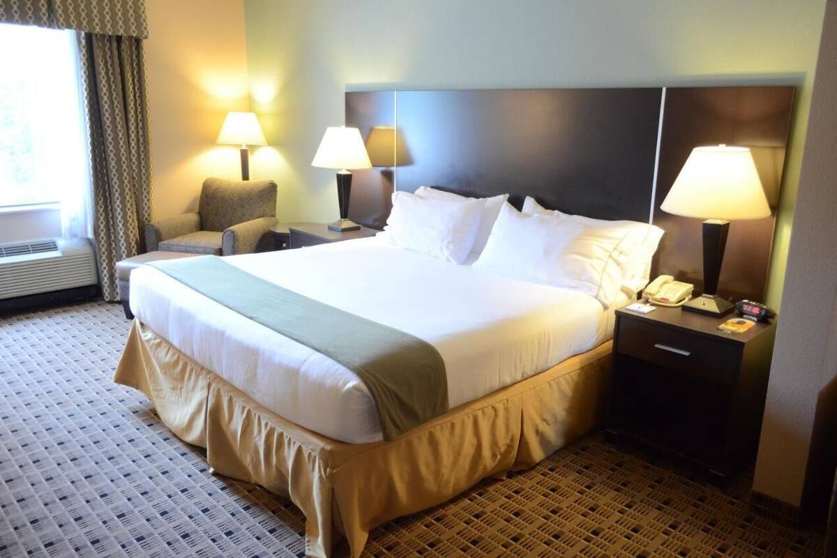 A hotel room with a large bed, two bedside lamps, a chair, a nightstand with a phone, and a window with curtains.
