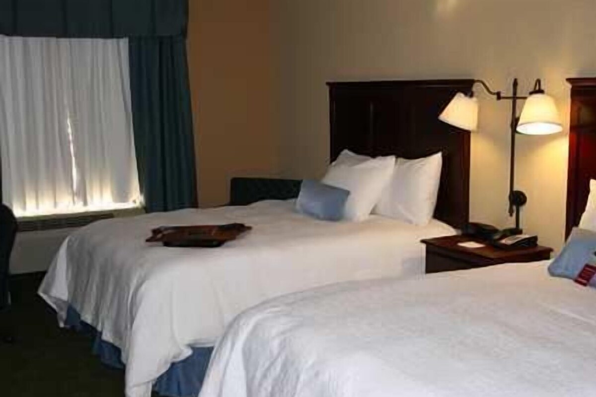 A hotel room with two beds, a desk, a lamp, and a window with curtains.