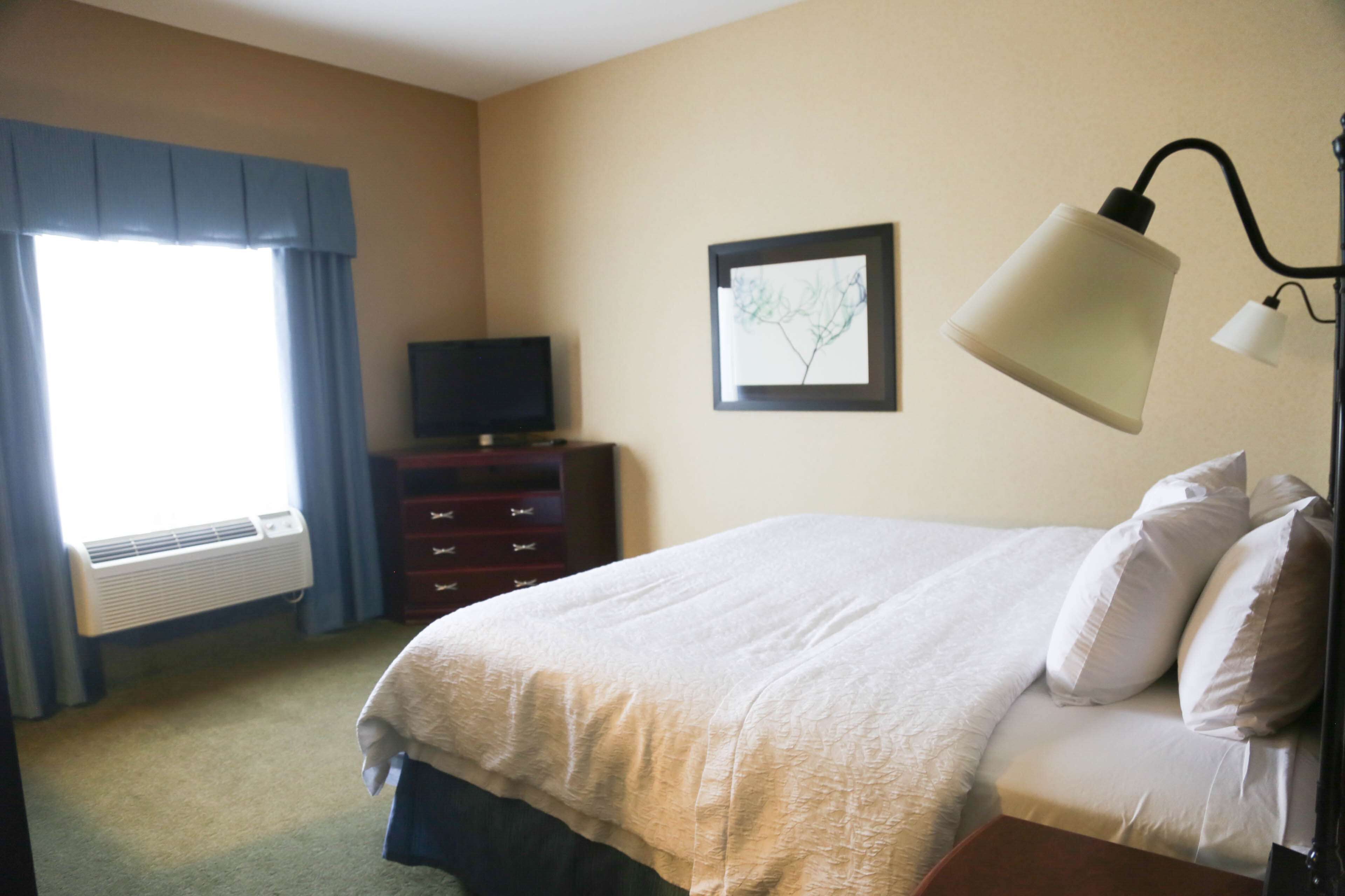 Habitación de hotel con cama, cómoda, televisor, un cuadro en la pared y ventana con cortinas.