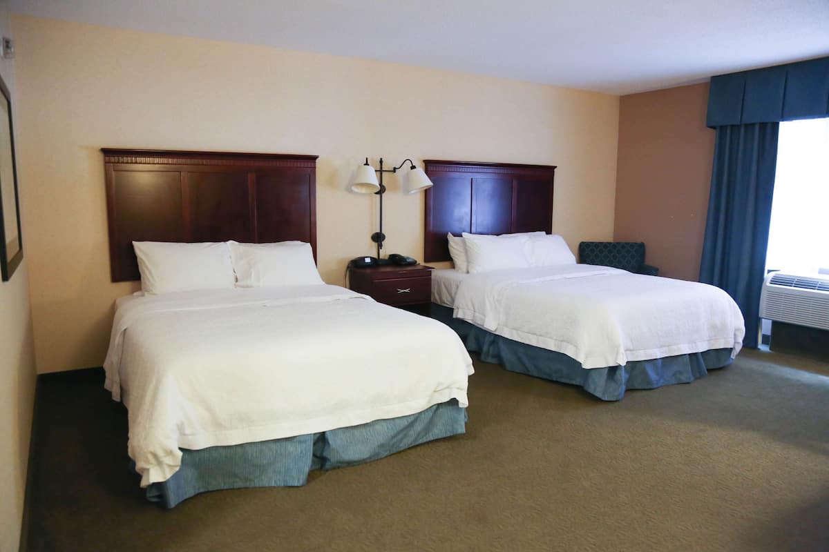 A hotel room with two beds, a nightstand, a lamp, and a window with curtains.