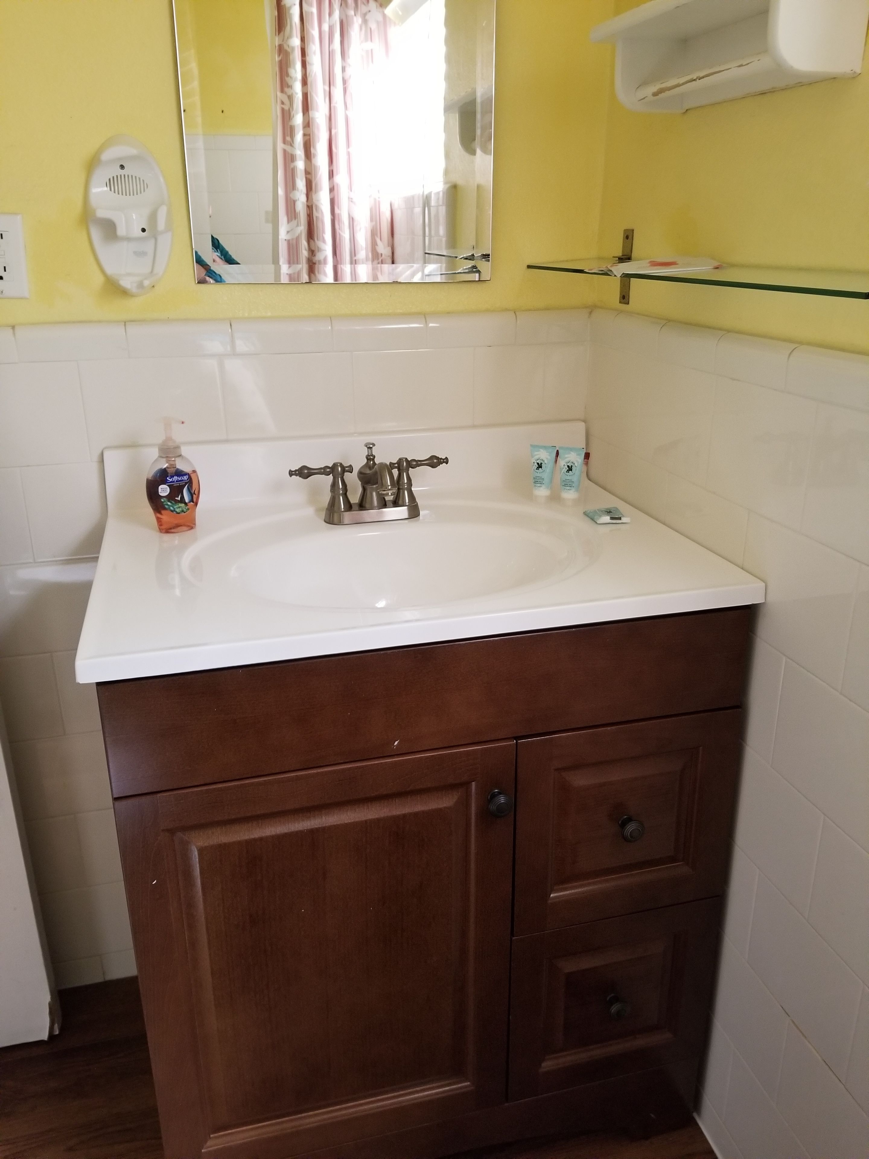 Un baño con un lavabo blanco, tocador de madera y un espejo. Hay un dispensador de jabón, un porta cepillos de dientes y un perchero para toallas en la pared.