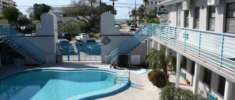 Une piscine bordée de bleu, un escalier menant à un bâtiment, et une vue sur la rue et les bâtiments en arrière-plan.
