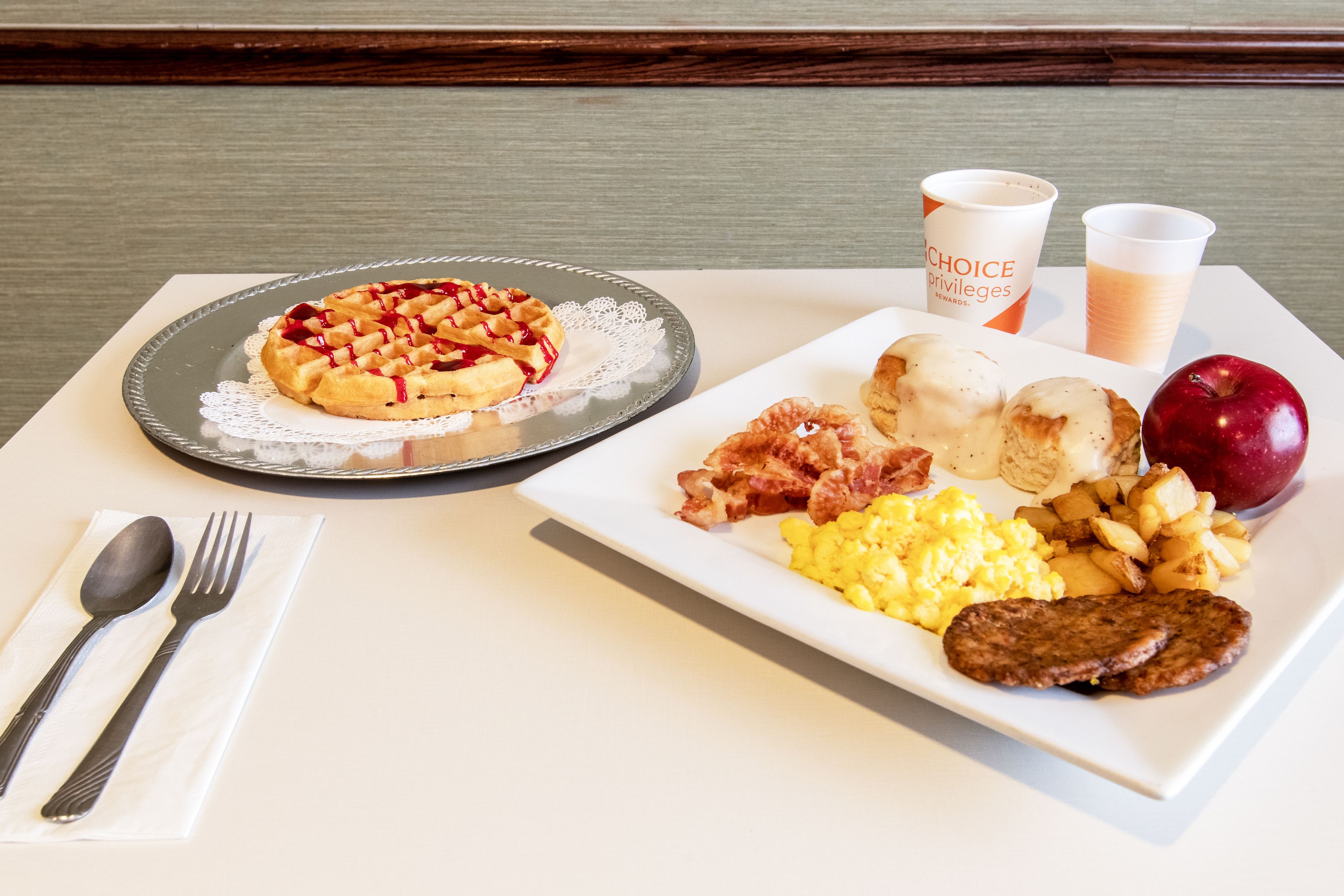A breakfast tray with a waffle, scrambled eggs, sausage, potatoes, and a red apple.