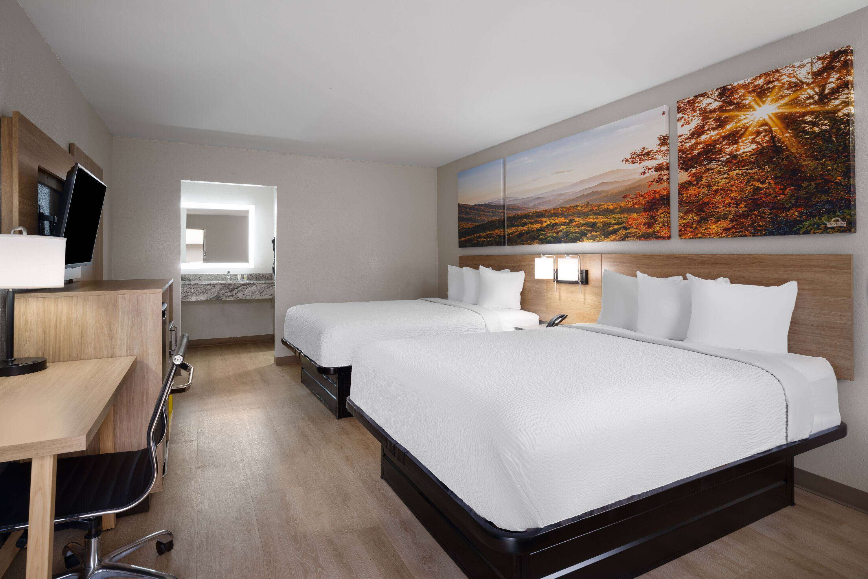 Habitación de hotel con dos camas, un escritorio con una computadora, una silla y un baño visible a través de una puerta abierta.