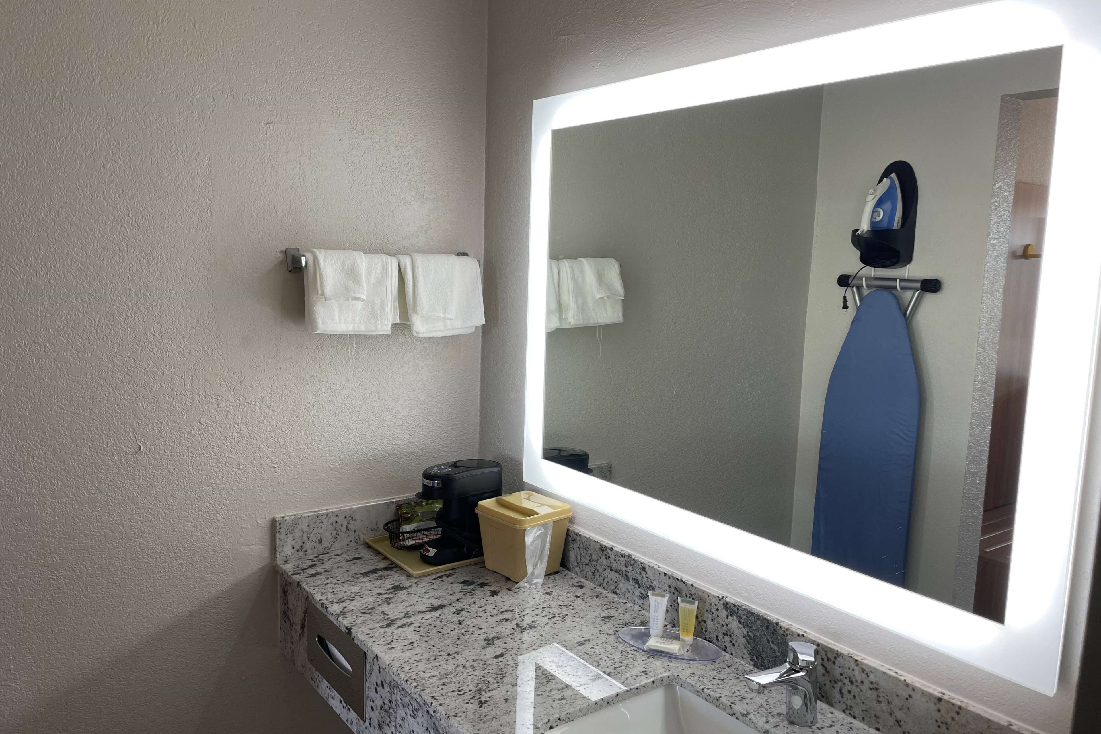 A bathroom with a large mirror, a towel rack with two towels, a coffee maker, a trash bin, and a small table with a glass of water.