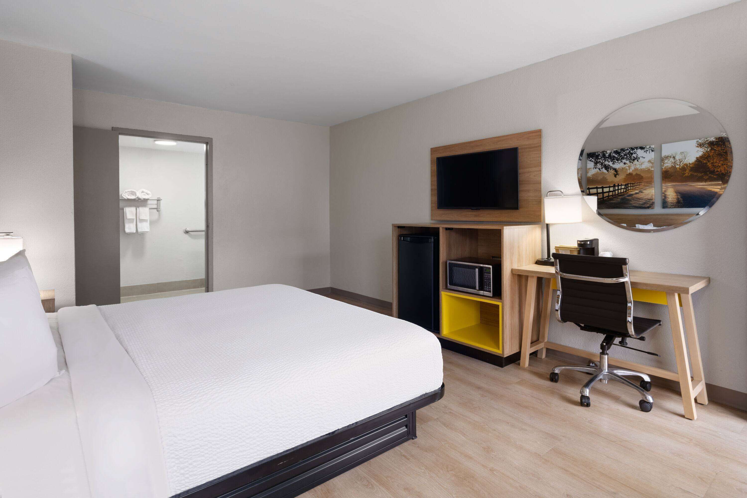 Habitación de hotel con cama, escritorio, computadora, televisor y espejo.