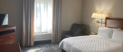A hotel room with a bed, a desk, a chair, a TV, a window with curtains, and a lamp.