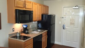 A compact kitchen with wooden cabinets, a black microwave, a refrigerator, and a coffee maker. - Candlewood Suites La Porte by IHG (La Porte)