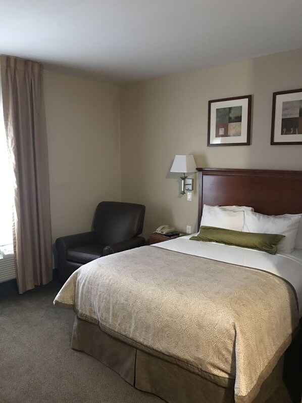 A hotel room with a large bed, a chair, a desk with a lamp, and two framed pictures on the wall.