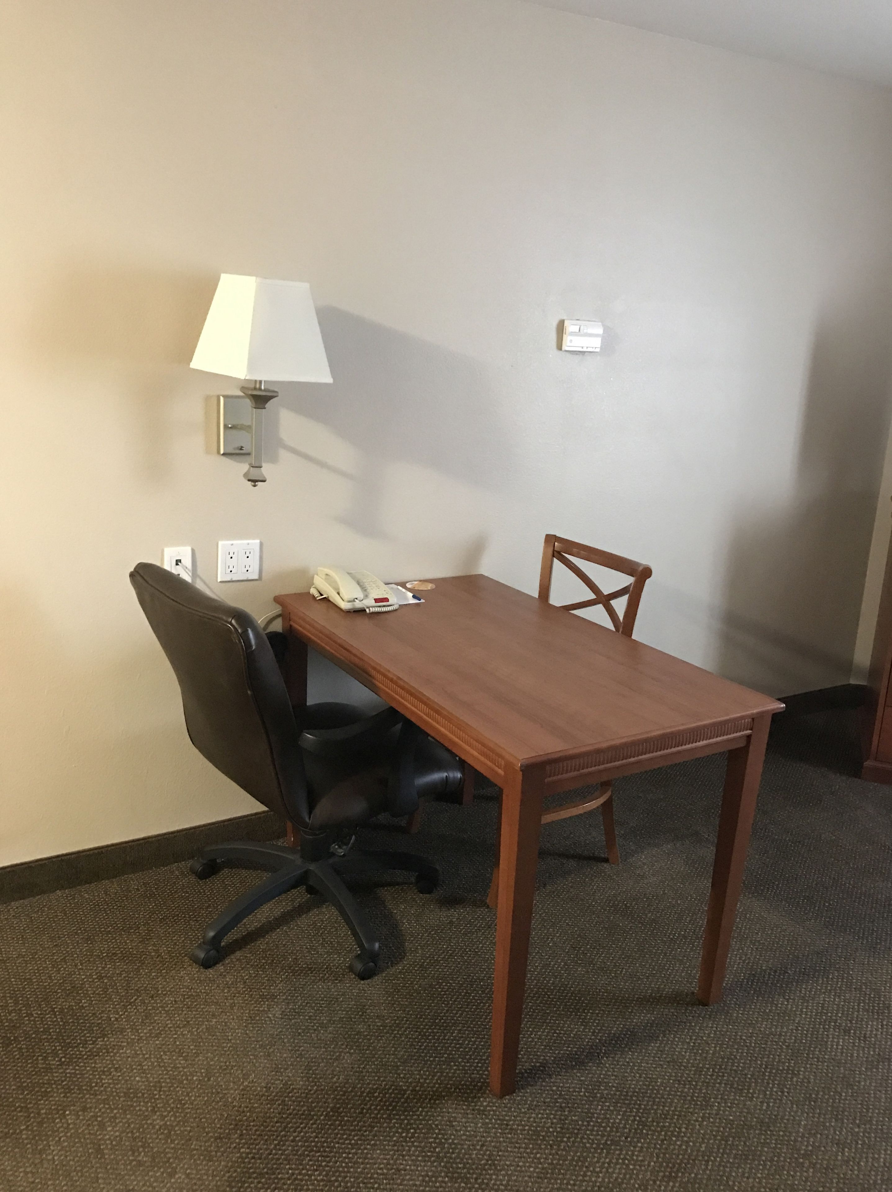 A small meeting room with a wooden table, two chairs, a desk lamp, a telephone, and a wall socket.