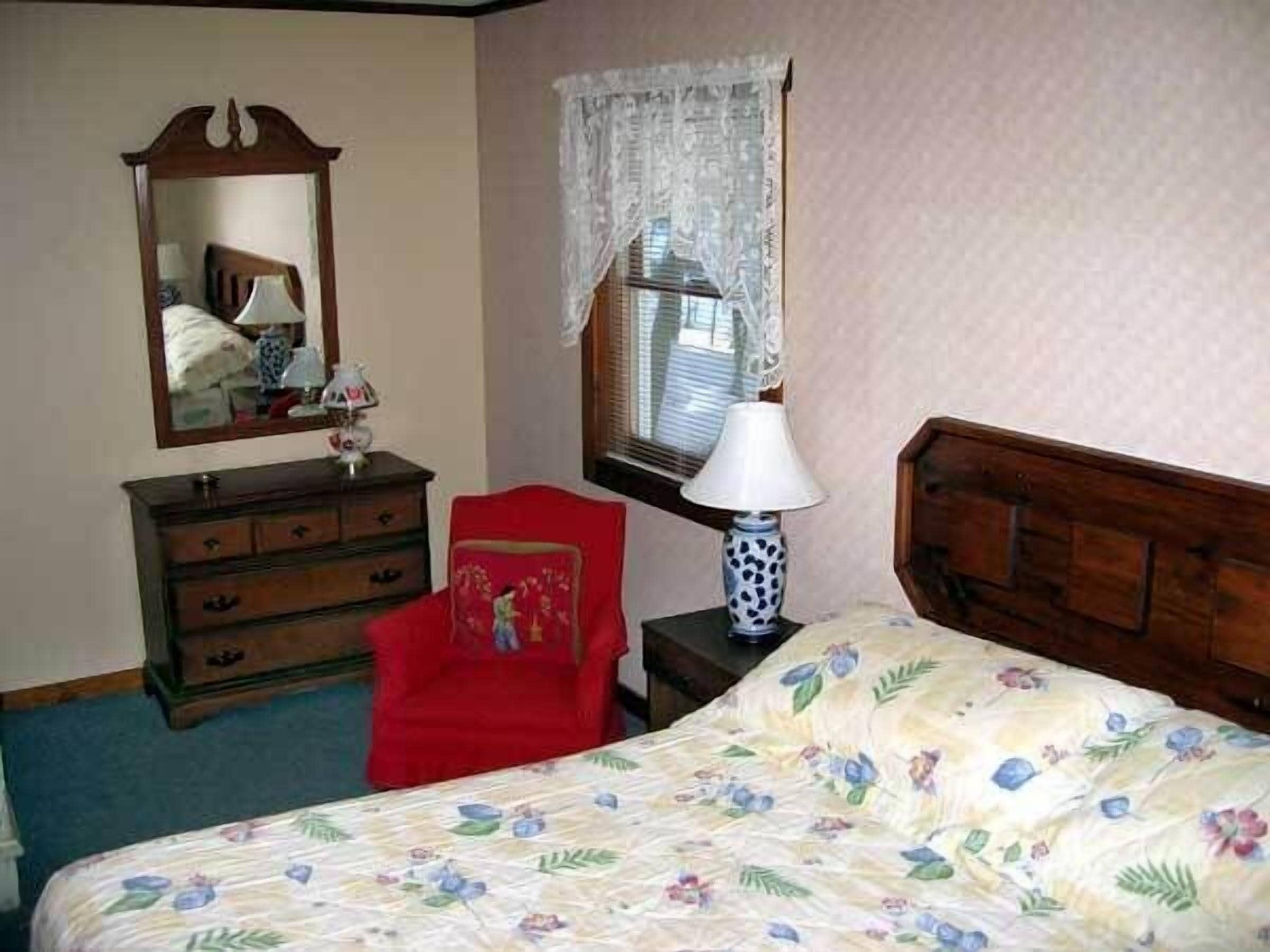 Een slaapkamer met een bed, een dressoir, een stoel, een nachtkastje, een lamp, een raam met kantgordijnen en een spiegel.