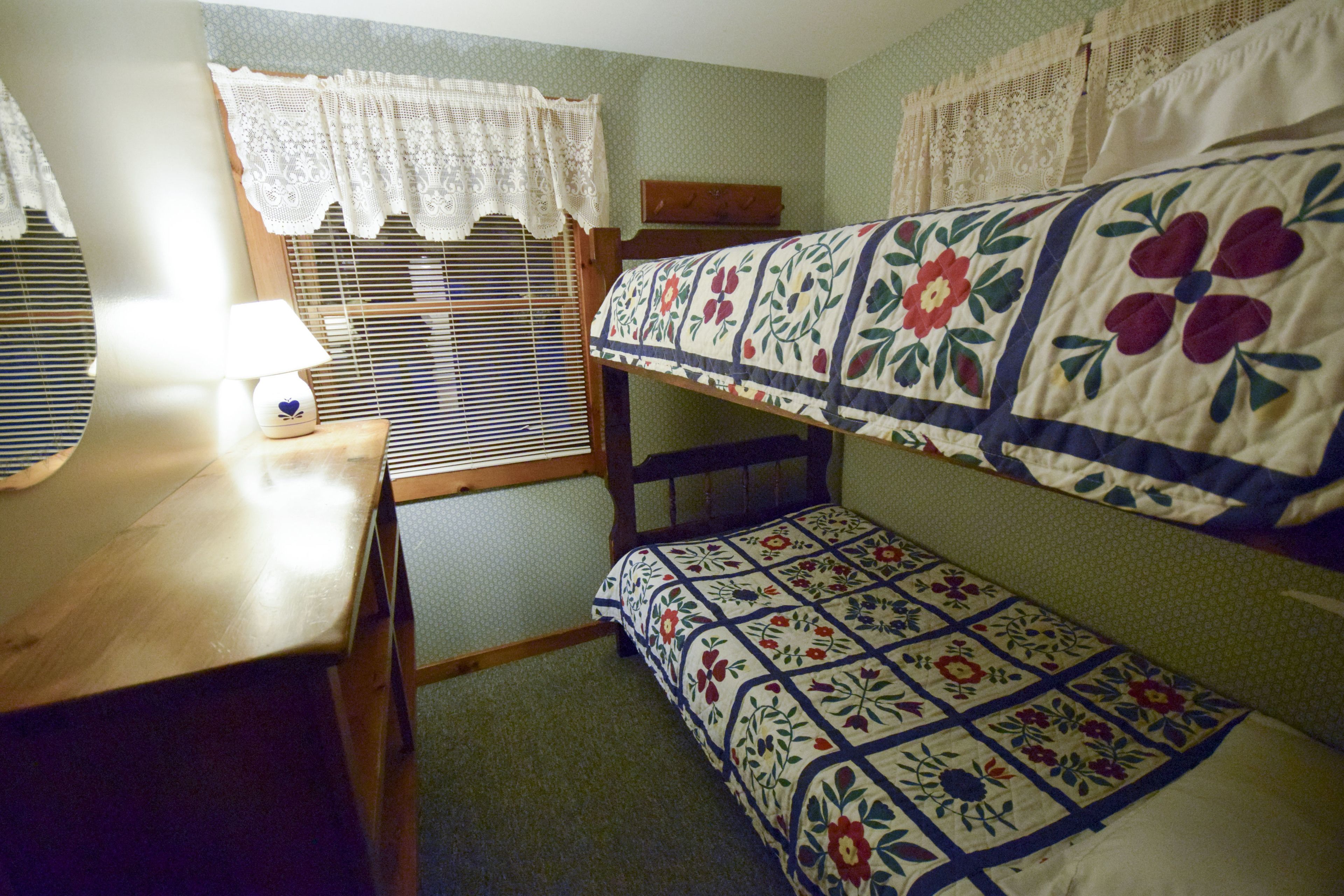A bunk bed with floral bedding, a wooden desk with a lamp, lace curtains, and a window with blinds.