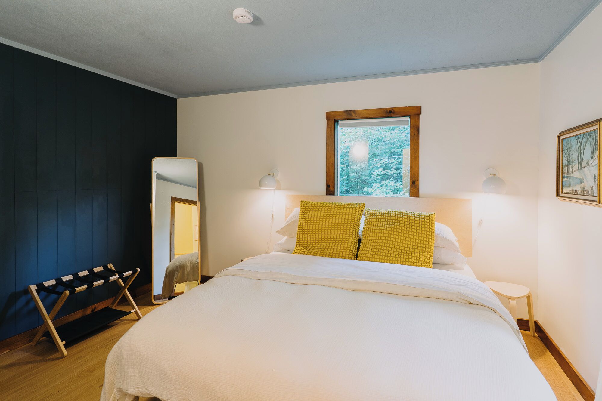 Une chambre à coucher avec un lit, un miroir, une fenêtre donnant sur des arbres et un tableau accroché au mur.