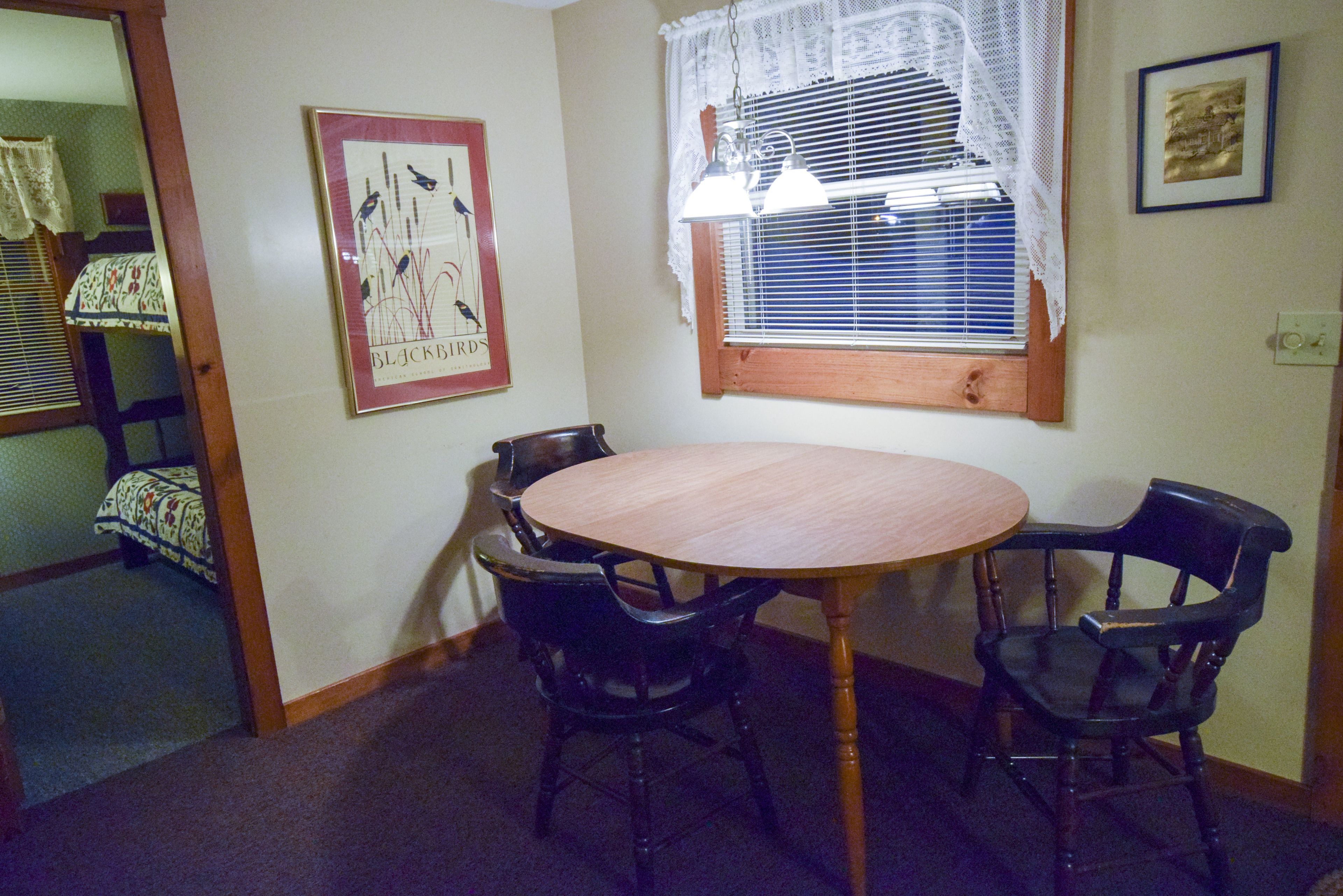 A dining area with a round table and four chairs, a window with blinds, and a framed picture on the wall.
