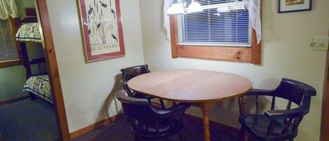 A dining area with a round table and four chairs, a window with blinds, and a framed picture on the wall.