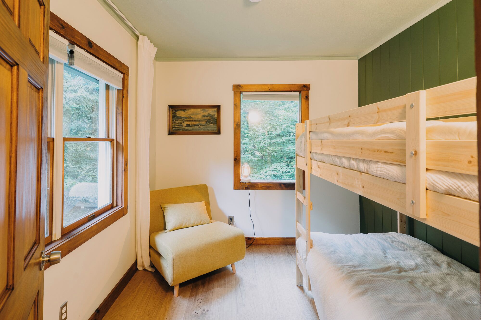 A room with a bunk bed, a single bed, a chair, a window with a view of trees, and a painting on the wall.