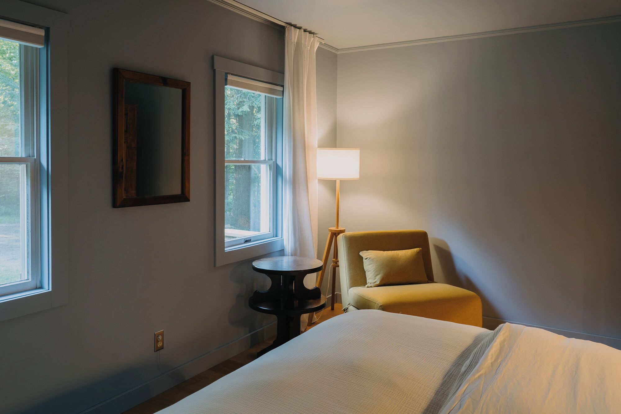 Una habitación de hotel con una cama, una silla, una lámpara y dos ventanas.