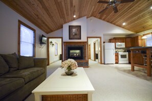 A spacious living room with a fireplace, a kitchen area, and a ceiling fan. - Stowe Cabins in the Woods (Waterbury Center)