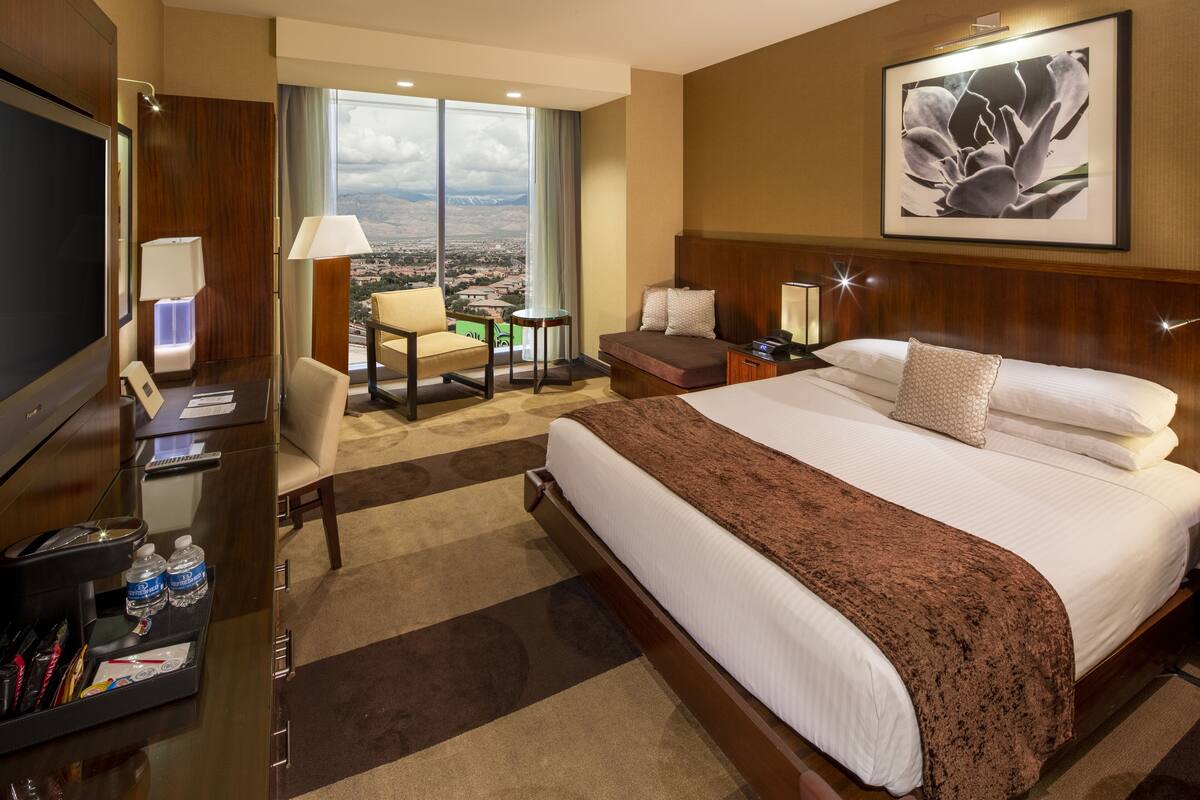 Hotelkamer met een groot bed, een bureau met een televisie, een stoel, een kleine tafel en uitzicht op de stad door grote ramen.