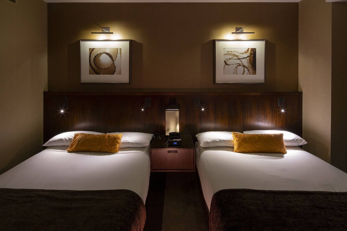 Een hotelkamer met twee bedden, elk voorzien van witte lakens en een bruin sierkussen. Boven de bedden hangen ingelijste kunstwerken en daartussen staat een klein tafeltje met een lamp.