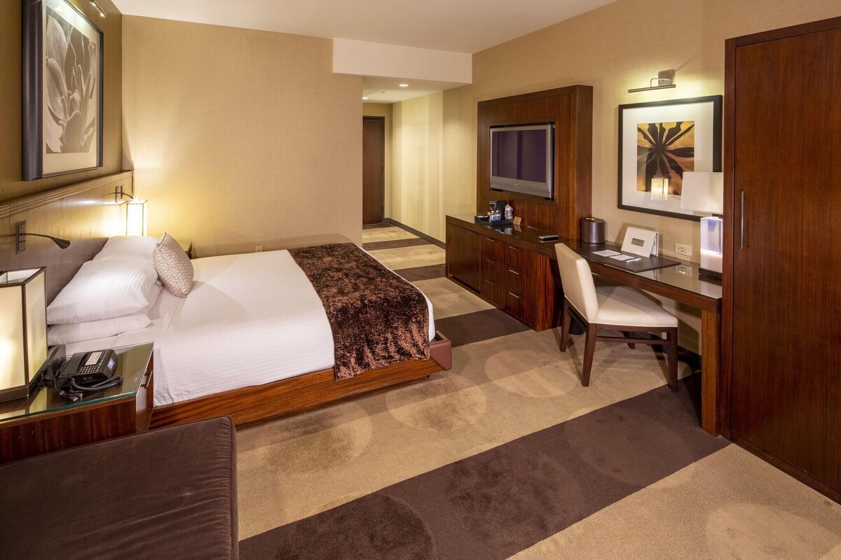 Quarto de hotel com cama, escrivaninha, cadeira, televisão e minibar.
