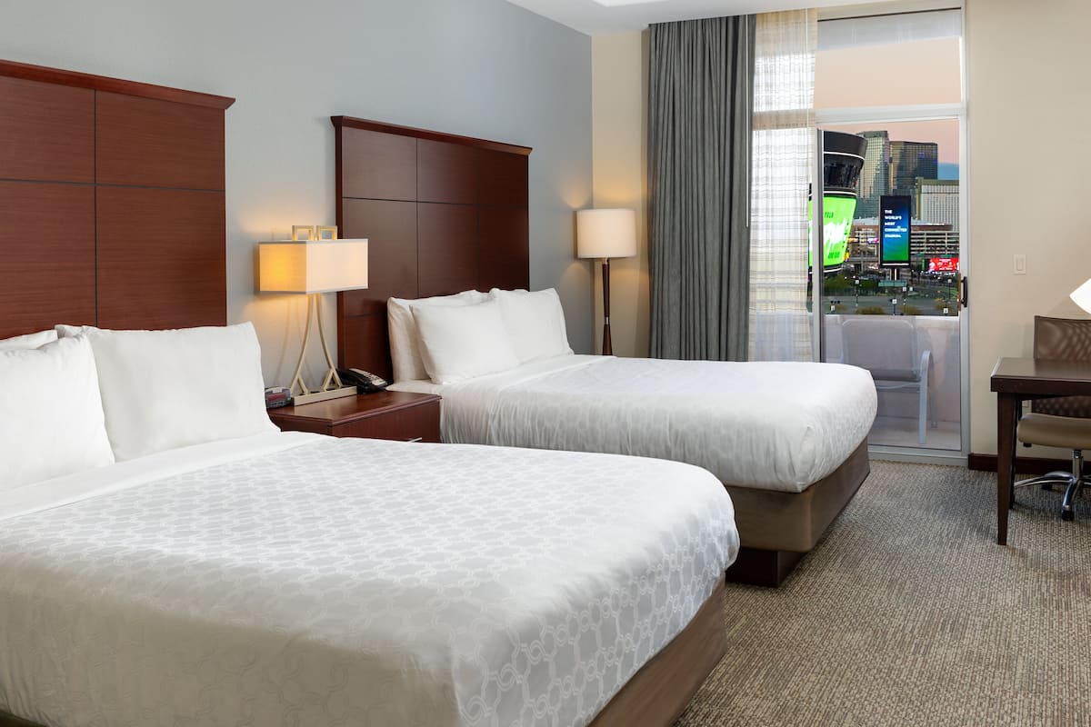 A hotel room with two beds, a desk, and a view of a cityscape through a large window.