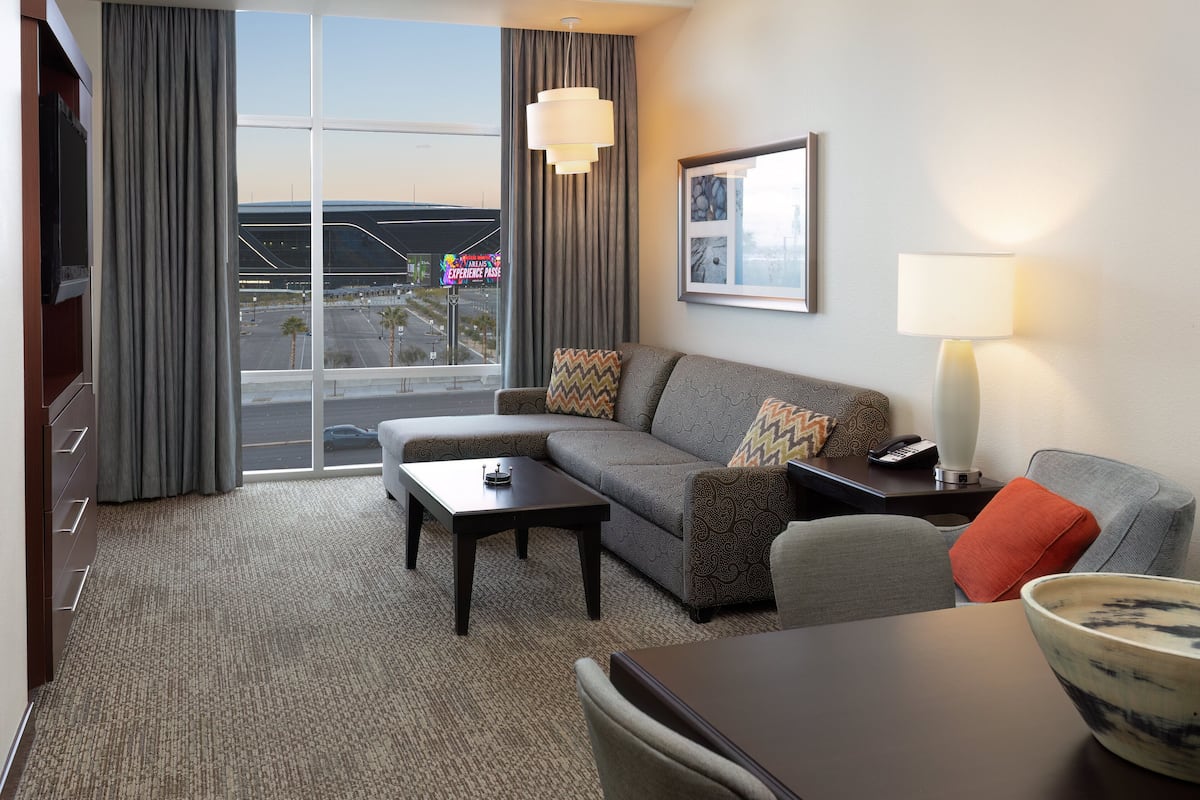 A modern hotel room with a large window, a sofa, a coffee table, a lamp, and a dining table.