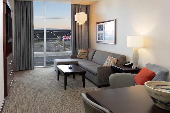 A modern hotel room with a large window, a sofa, a coffee table, a lamp, and a dining table.