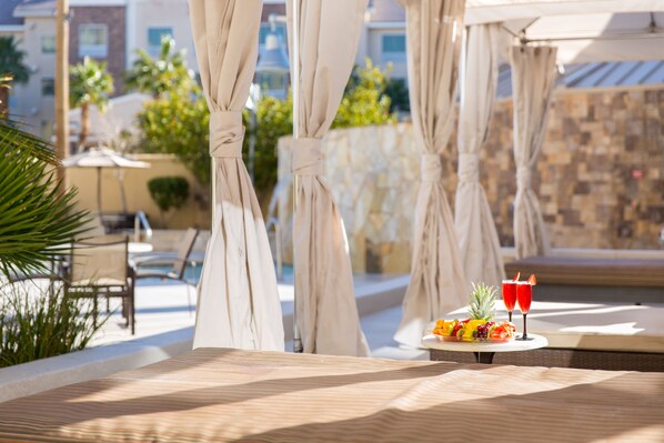 A sunlit outdoor seating area with a table set for two, featuring a fruit platter and two drinks.