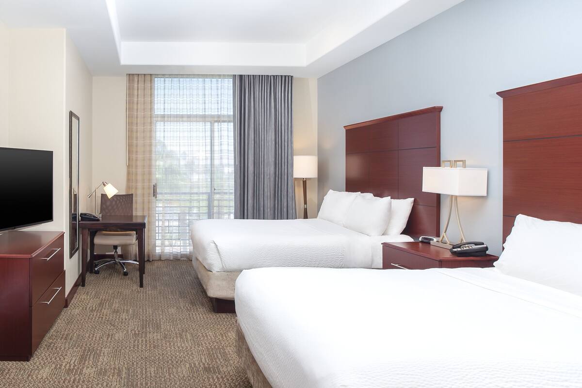 A hotel room with two beds, a desk, a TV, and a window with curtains.