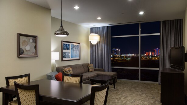 A hotel room with a dining area, a sofa, a TV, and a large window with a city view at night.