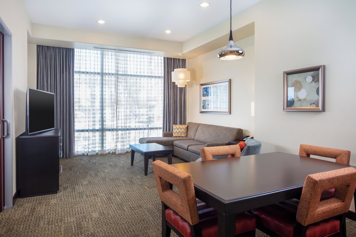 A hotel room with a sofa, a dining table with chairs, a flat-screen TV, and a large window with curtains.